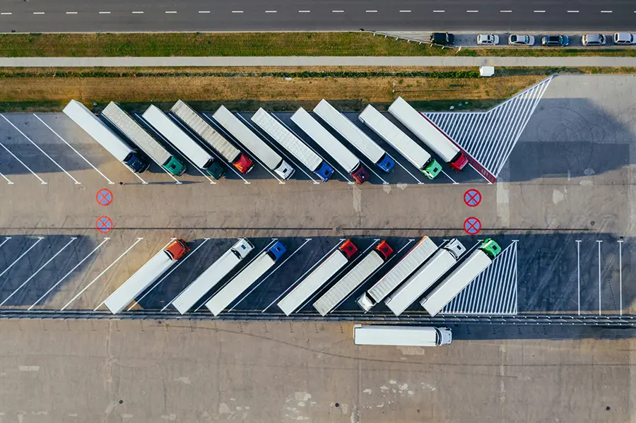 Aerial view of a parking area with multiple semi-trailer trucks parked diagonally in designated spaces. The lot includes marked no-parking zones and a clearly visible road at the top of the image with cars driving by. The scene is well-organized, indicating a logistics or transport hub.